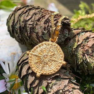 May include: Gold pendant necklace with a circular design featuring a sunburst pattern. The pendant is on a chain and resting on a piece of tree bark.