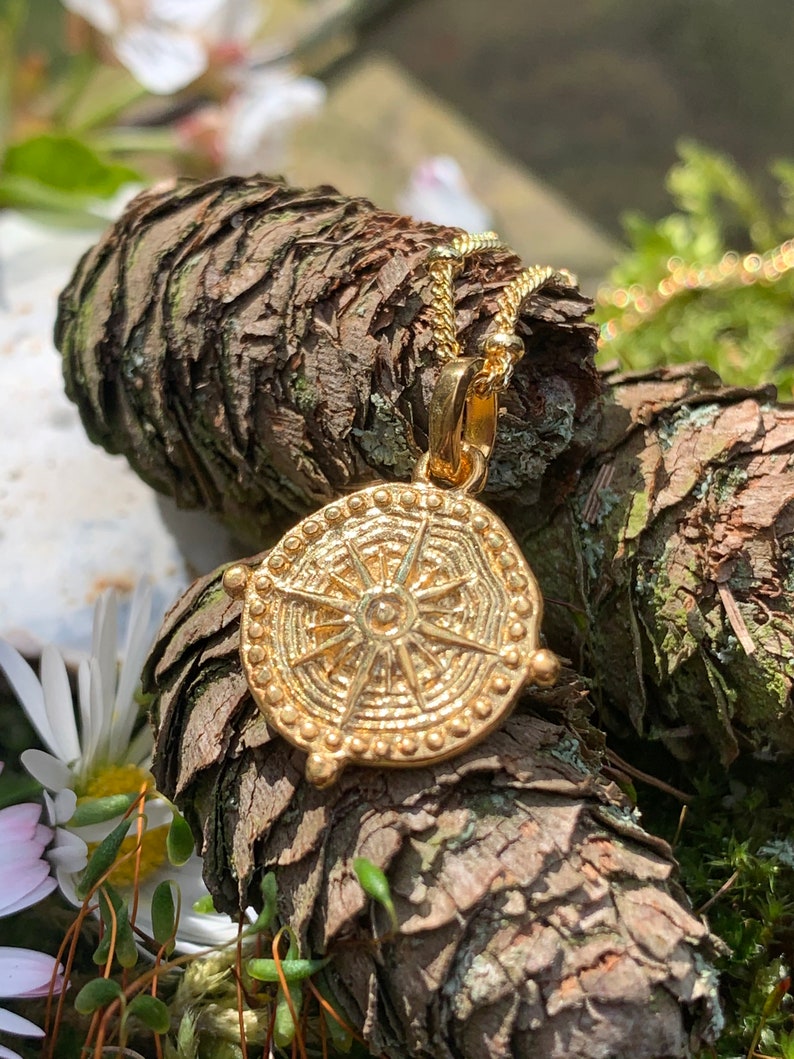 May include: Gold pendant necklace with a circular design featuring a sunburst pattern. The pendant is on a chain and resting on a piece of tree bark.