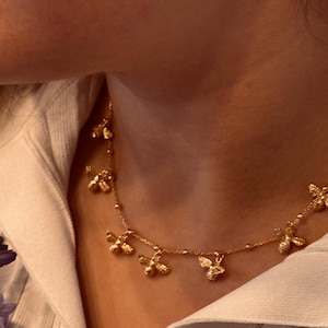 May include: A gold necklace with multiple bee pendants. The necklace is worn against a white collared shirt. The bees are detailed and appear to be made of metal. The necklace is delicate and elegant.