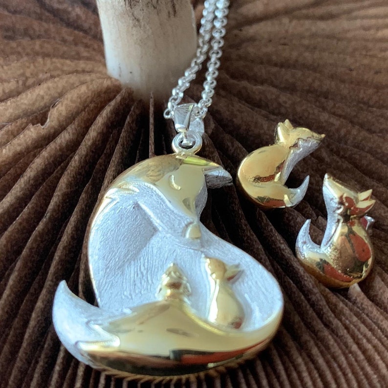 May include: A silver and gold fox pendant necklace with matching fox stud earrings. The pendant features a mother fox with two smaller foxes. The jewelry has a detailed design and is set against a brown background.