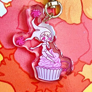 Può includere: Un portachiavi a forma di cupcake rosa e bianco con un personaggio dei cartoni animati seduto sopra. Il personaggio ha i capelli rosa, un vestito bianco e una coda rosa. Il portachiavi ha una chiusura dorata a forma di cuore.