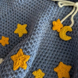 Puede incluir: Una capa con capucha de color azul hecha a crochet con adornos de estrellas amarillas. La capa tiene un ribete de crochet blanco y un cord&oacute;n blanco.