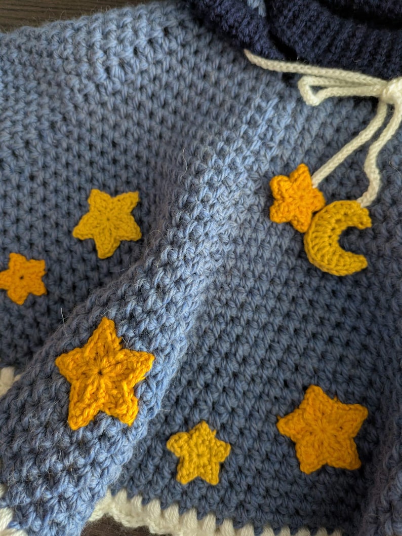 Puede incluir: Una capa con capucha de color azul hecha a crochet con adornos de estrellas amarillas. La capa tiene un ribete de crochet blanco y un cord&oacute;n blanco.