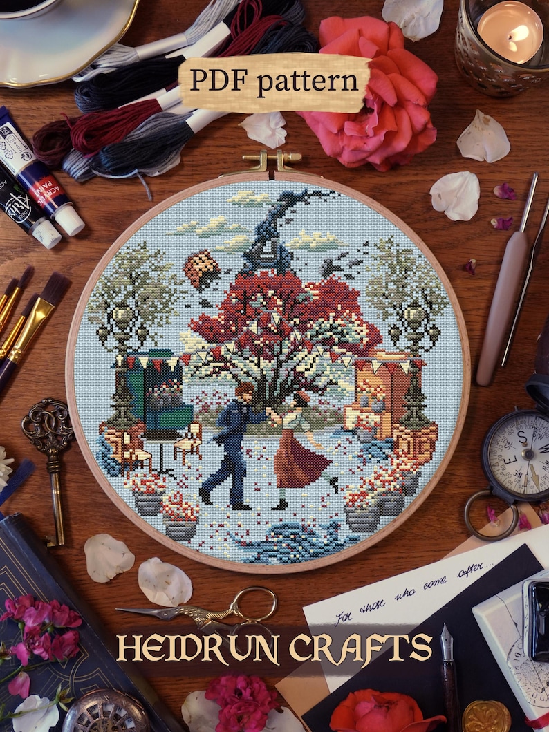 Finished embroidery displayed in a loop of the Gustave and Sophie cross stitch pattern. Inspired by a dance scene in Clair Obscur : Expedition 33 game of the year. The couple dances in front of the red and white tree in Lumière with the stalls and bent Eiffel Tower. Red and white petals, brushes, old books and stationary frame the composition. PDF pattern available at Heidrun Crafts shop.