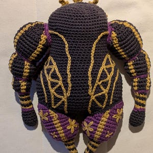 Pu&ograve; includere: Bambola all'uncinetto fatta a mano in blu scuro, oro e viola. La bambola ha una corona, maniche a sbuffo e cuciture dorate decorative. Le gambe e i piedi sono dettagliati con accenti viola e oro, su una superficie bianca.