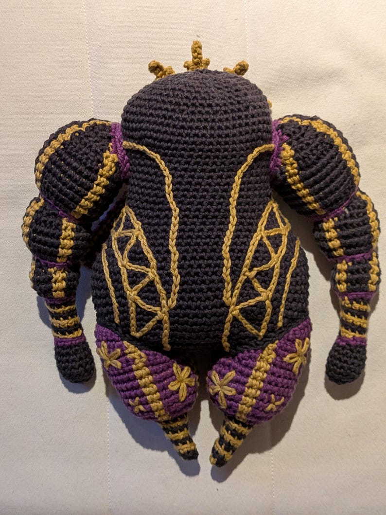 Pu&ograve; includere: Bambola all'uncinetto fatta a mano in blu scuro, oro e viola. La bambola ha una corona, maniche a sbuffo e cuciture dorate decorative. Le gambe e i piedi sono dettagliati con accenti viola e oro, su una superficie bianca.