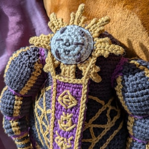 Pu&ograve; includere: Bambola all'uncinetto fatta a mano con un dettaglio a forma di sole. La bambola ha una faccia sorridente e indossa un abito viola scuro e oro. Il design include motivi dettagliati, evidenziando l'artigianato dell'articolo fatto a mano.
