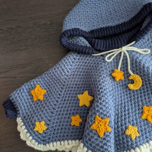 Puede incluir: Un poncho azul de ganchillo con capucha, ribete de ganchillo blanco y aplicaciones de estrellas y luna creciente amarillas.