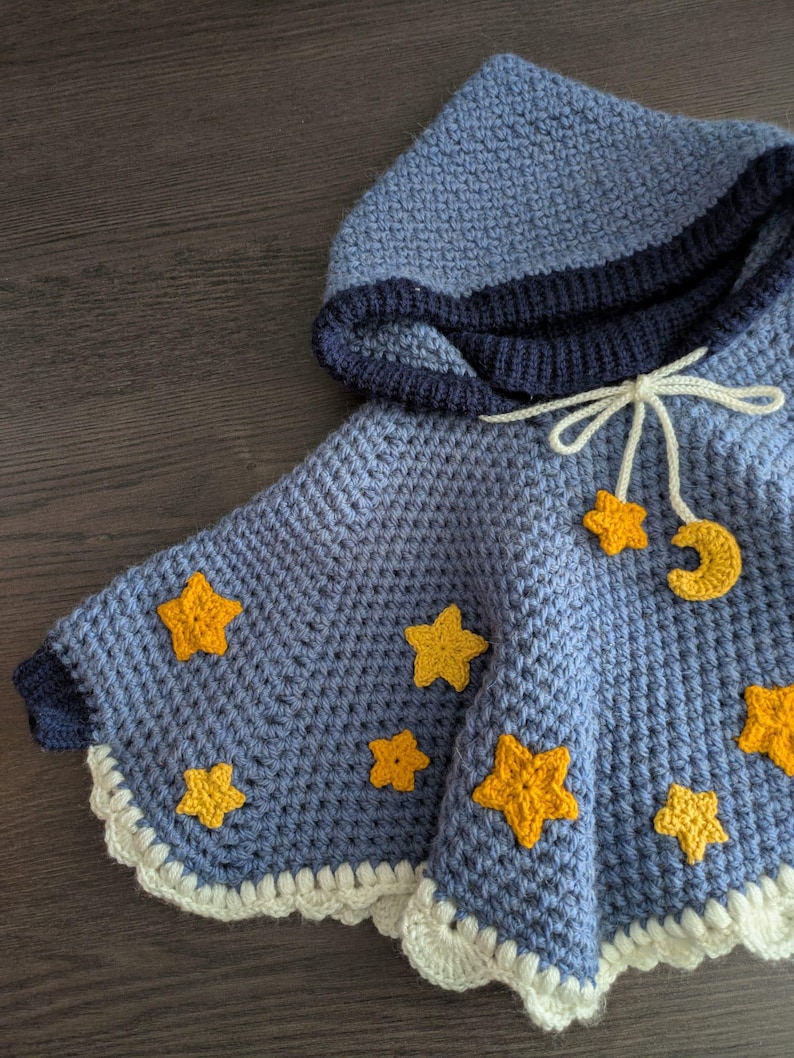Puede incluir: Un poncho azul de ganchillo con capucha, ribete de ganchillo blanco y aplicaciones de estrellas y luna creciente amarillas.
