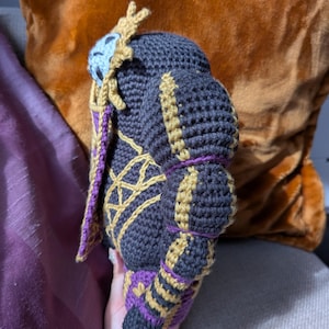 Pu&ograve; includere: Bambola all'uncinetto fatta a mano nelle tonalit&agrave; del grigio scuro, oro e viola. La bambola presenta cuciture intricate e un design dettagliato, con un abbellimento a forma di corona sulla testa. La bambola &egrave; tenuta contro uno sfondo di un cuscino marrone e viola.