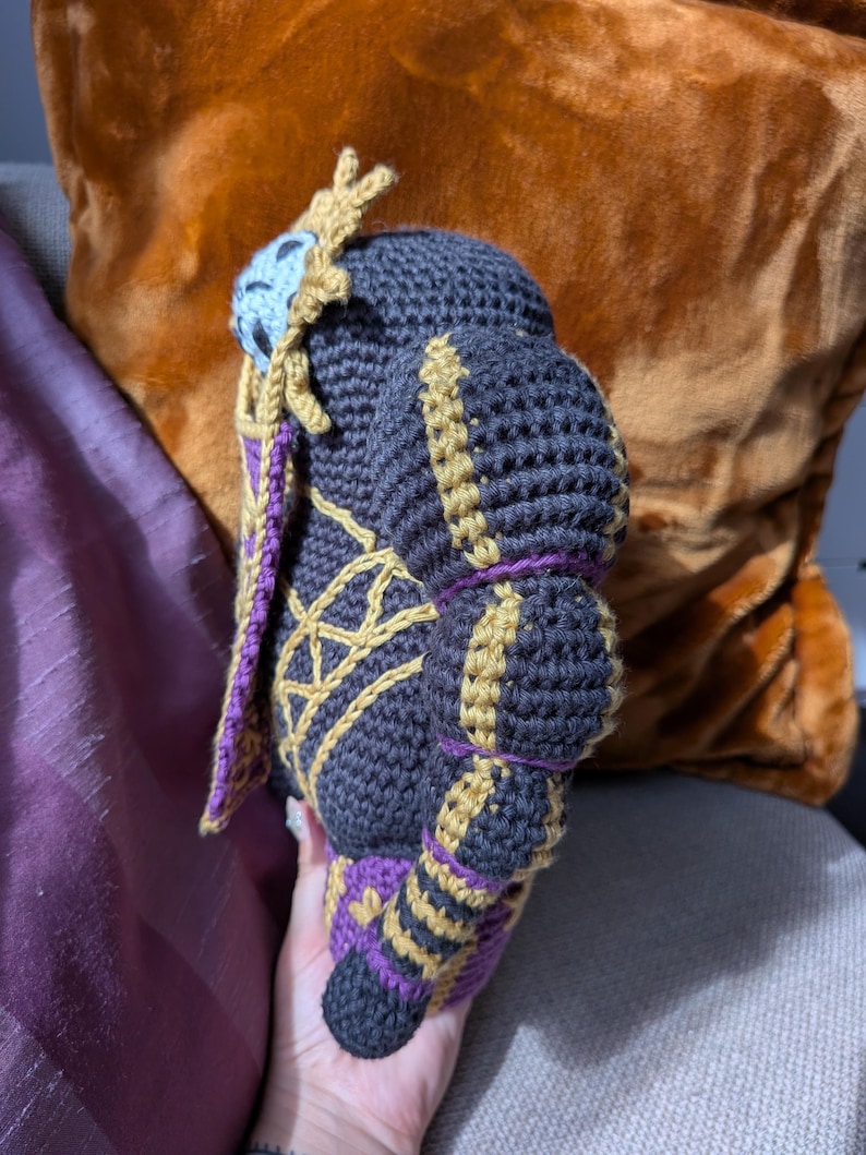 Pu&ograve; includere: Bambola all'uncinetto fatta a mano nelle tonalit&agrave; del grigio scuro, oro e viola. La bambola presenta cuciture intricate e un design dettagliato, con un abbellimento a forma di corona sulla testa. La bambola &egrave; tenuta contro uno sfondo di un cuscino marrone e viola.
