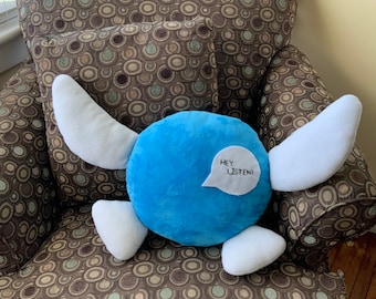 May include: A blue plush toy with white wings and a speech bubble that says "Hey Listen!" The toy is sitting on a brown patterned armchair.