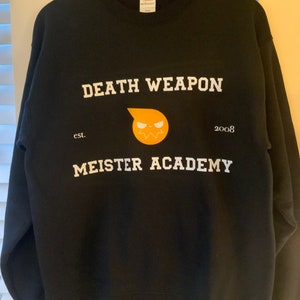 Op de afbeelding: Zwarte sweatshirt met de witte tekst "DEATH WEAPON MEISTER ACADEMY" en een gele en oranje afbeelding. De sweatshirt heeft ook de tekst "est. 2008". Het hangt aan een houten kleerhanger.