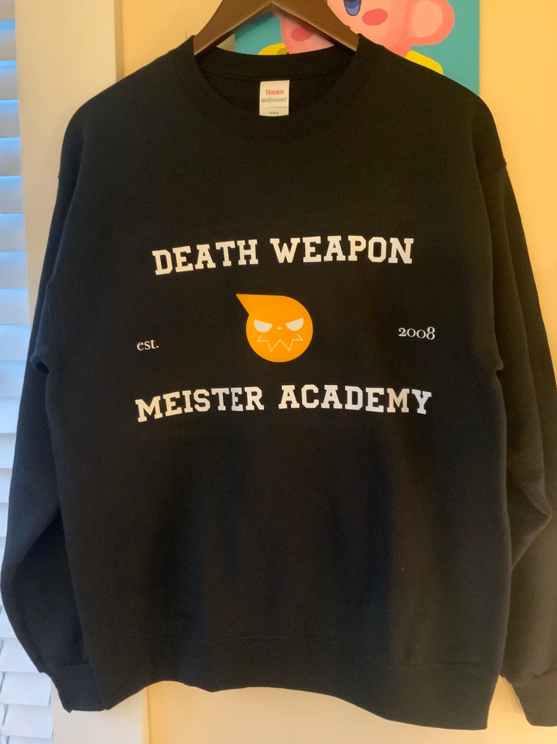 Op de afbeelding: Zwarte sweatshirt met de witte tekst "DEATH WEAPON MEISTER ACADEMY" en een gele en oranje afbeelding. De sweatshirt heeft ook de tekst "est. 2008". Het hangt aan een houten kleerhanger.
