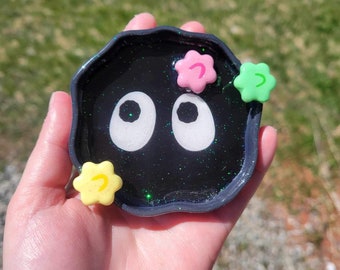 May include: A black resin coaster with glitter and white eyes. It has three small flower-shaped charms in pink, green, and yellow.