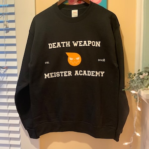 Op de afbeelding: Zwart sweatshirt met witte tekst "DEATH WEAPON MEISTER ACADEMY", een oranje afbeelding en het jaar "2008". Het sweatshirt hangt aan een houten kleerhanger.