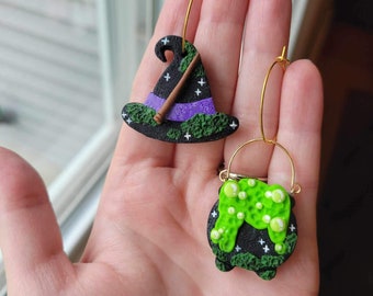 May include: A pair of witch-themed earrings. One earring features a black witch hat with a purple band and a brown broomstick. The other earring is a green cauldron with a black rim and white stars. Both earrings have a gold hoop.