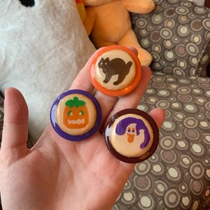 May include: Three Halloween-themed pinback buttons. One button features a black cat on an orange background, another has a jack-o'-lantern on a purple background, and the last has a ghost on a brown background. The buttons are round and have a glossy finish.