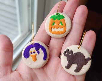 May include: Three Halloween-themed clay ornaments with gold hoops. The ornaments are a purple ghost with an orange face, a brown cat, and an orange pumpkin with a green stem and a jack-o'-lantern face.