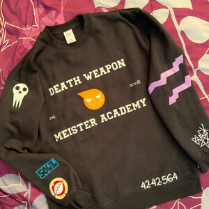 Op de afbeelding: Zwart sweatshirt met witte tekst "DEATH WEAPON MEISTER ACADEMY" en "2008". Het sweatshirt heeft een cartoon schedel en een cartoon zon met een gezicht. De mouwen hebben paarse en witte ontwerpen en patches.
