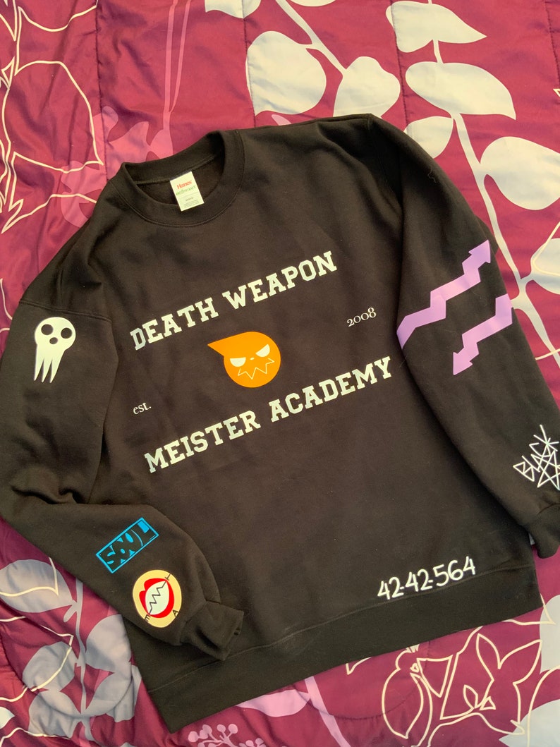 Op de afbeelding: Zwart sweatshirt met witte tekst "DEATH WEAPON MEISTER ACADEMY" en "2008". Het sweatshirt heeft een cartoon schedel en een cartoon zon met een gezicht. De mouwen hebben paarse en witte ontwerpen en patches.