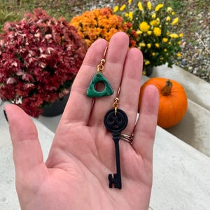 May include: A pair of earrings featuring a green triangle charm and a black key charm. The triangle charm has a circle cut out of the centre. The key charm has a black button on the top.