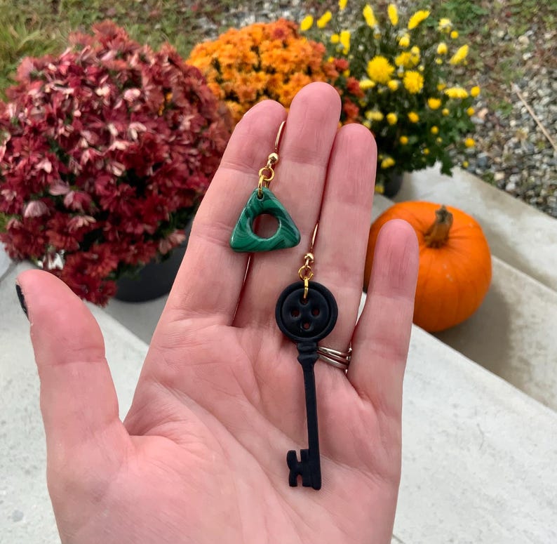 May include: A pair of earrings featuring a green triangle charm and a black key charm. The triangle charm has a circle cut out of the centre. The key charm has a black button on the top.