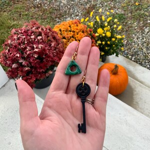 May include: A pair of dangle earrings. One earring is a green triangle with a hole in the centre. The other earring is a black key with a skull-shaped head.
