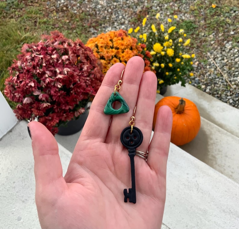 May include: A pair of dangle earrings. One earring is a green triangle with a hole in the centre. The other earring is a black key with a skull-shaped head.
