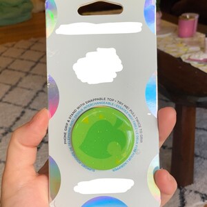 May include: A green and yellow phone grip with a leaf design and glitter. The grip is attached to a white card with the text "SUPPORT-POIGNÉE DE TÉLÉPHONE AVEC DESSUS INTERCHANGEABLE - ESSAYEZ-MOI! TIREZ-LE DEUX FOIS POUR L'AGRIPPER."