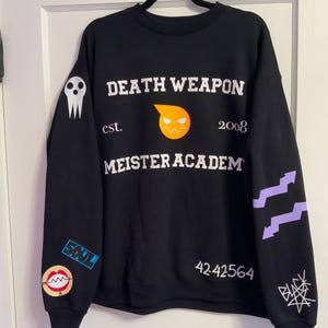 Op de afbeelding: Zwart sweatshirt met witte tekst "DEATH WEAPON MEISTER ACADEM" en "est. 2008". Het ontwerp bevat een schedelafbeelding, een gestileerde oranje gezichtsafbeelding en extra afbeeldingen en tekst op de mouwen. Het sweatshirt hangt aan een zwarte hanger.