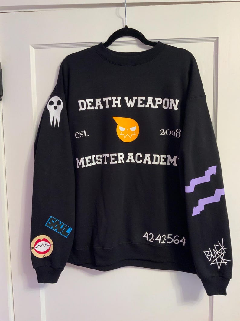 Op de afbeelding: Zwart sweatshirt met witte tekst "DEATH WEAPON MEISTER ACADEM" en "est. 2008". Het ontwerp bevat een schedelafbeelding, een gestileerde oranje gezichtsafbeelding en extra afbeeldingen en tekst op de mouwen. Het sweatshirt hangt aan een zwarte hanger.