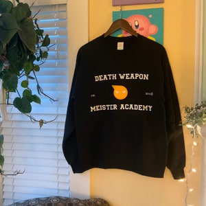 Op de afbeelding: Een zwart sweatshirt met witte tekst "DEATH WEAPON MEISTER ACADEMY" en een geel en oranje afbeelding. Het sweatshirt hangt aan een houten hanger tegen een gele muur. De tekst bevat het jaar "2008".