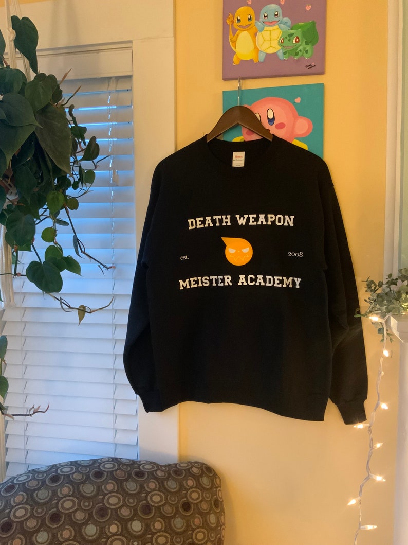 Op de afbeelding: Een zwart sweatshirt met witte tekst "DEATH WEAPON MEISTER ACADEMY" en een geel en oranje afbeelding. Het sweatshirt hangt aan een houten hanger tegen een gele muur. De tekst bevat het jaar "2008".