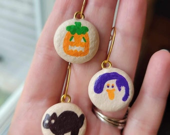 May include: Three Halloween-themed earrings with gold hoops. The earrings are painted on round, white, wooden bases. One earring features a black cat, one features a purple ghost, and one features an orange pumpkin.