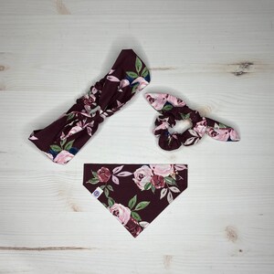 May include: A set of three burgundy floral hair accessories. The set includes a headband, a scrunchie, and a bandana. All three accessories feature a floral pattern with pink, white, and blue flowers on a burgundy background.