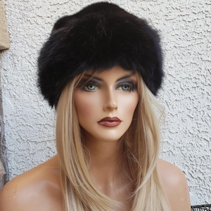 May include: A black fur hat with a rounded crown. The hat is worn by a mannequin with blonde hair and blue eyes. The mannequin is in front of a textured, off-white wall.