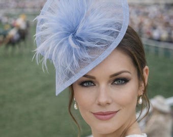 Può includere: Un cappello fascinator azzurro con base in rete e dettagli di piume. La persona indossa il cappello a un evento all'aperto, con uno sfondo sfocato di un ippodromo.