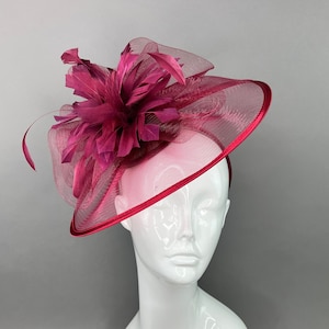 May include: A burgundy fascinator with a large feather flower and a net veil. The fascinator is on a white mannequin head.