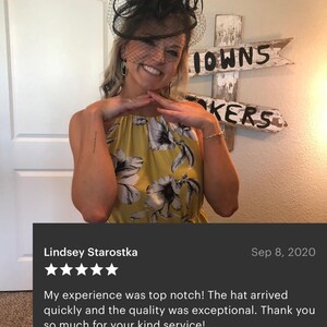 May include: A black fascinator hat featuring a decorative arrangement of feathers and a fine mesh veil that covers the upper face. The headpiece is paired with a sleeveless dress in a mustard yellow color, adorned with a large black and white floral print.