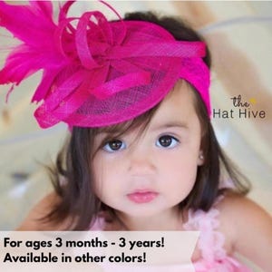 May include: A bright pink headband with a large, decorative flower and feather accents. The headband is worn by a young child. The text "the Hat Hive" is visible in the upper right corner. The text "For ages 3 months - 3 years! Available in other colors!" is at the bottom.