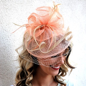 May include: A peach-colored fascinator with a veil. The headpiece features a layered design with mesh and feather accents. The veil is a fine, off-white netting. The overall style is elegant and suitable for formal events.