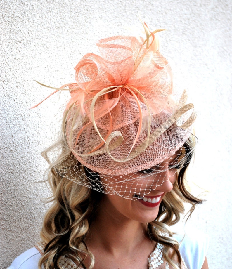 May include: A peach-colored fascinator with a veil. The headpiece features a layered design with mesh and feather accents. The veil is a fine, off-white netting. The overall style is elegant and suitable for formal events.