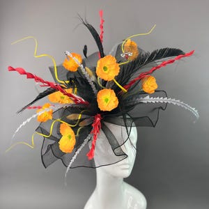 May include: A fascinator hat featuring black tulle netting, vibrant orange poppy flowers, long black feathers, red spiral accents, and thin yellow wire stems. A small decorative bee embellishment is attached to the black mesh fabric.