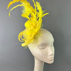 May include: A bright yellow fascinator with a mix of feathers and fabric accents. The headpiece is mounted on a mannequin head, showcasing its design. The feathers are arranged to create a dynamic, eye-catching look.