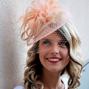 May include: A peach-colored fascinator with a delicate veil and feather accents. The headpiece is made of sheer fabric and is adorned with a bow. This elegant accessory is perfect for special occasions and events.