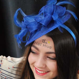 May include: A vibrant blue headpiece with a decorative bow and feather accents. The headpiece is attached to a thin headband. The text "the Hat Hive" is visible on the forehead. This accessory is suitable for special occasions.