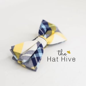 May include: A handmade bow tie with a plaid pattern in yellow, navy blue, and white. The bow tie is knotted in the center. The text "the Hat Hive" is printed in black.