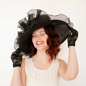 May include: A woman wearing a black lace hat with a large brim and black lace gloves. She is wearing a white lace dress and has her eyes closed.