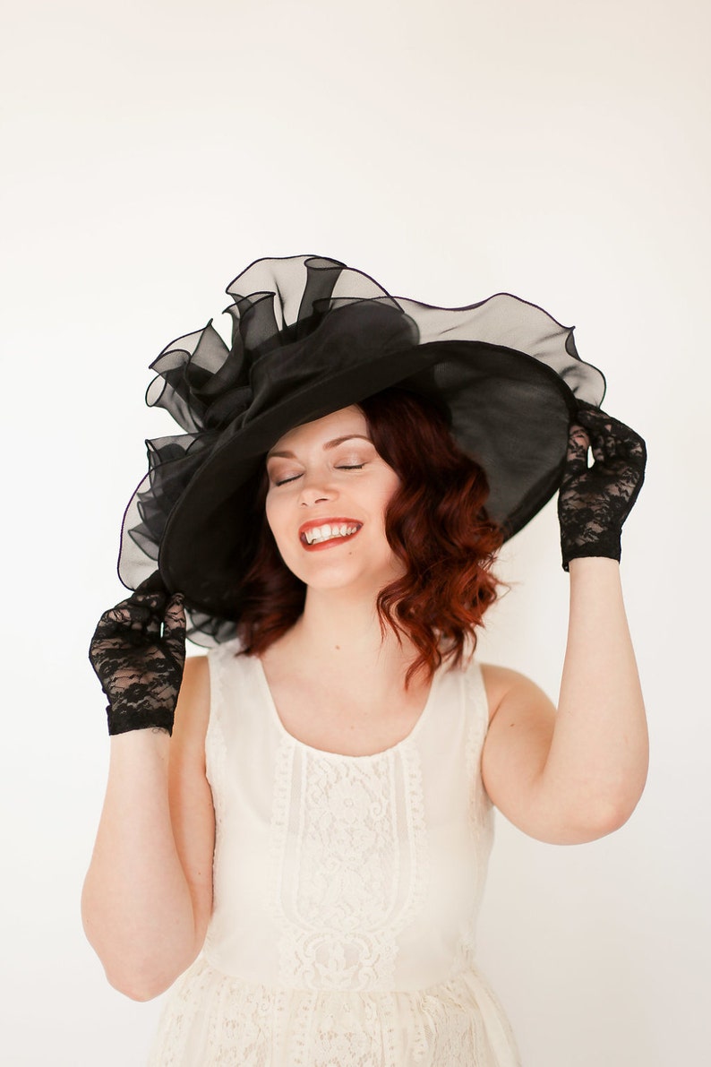 May include: A woman wearing a black lace hat with a large brim and black lace gloves. She is wearing a white lace dress and has her eyes closed.